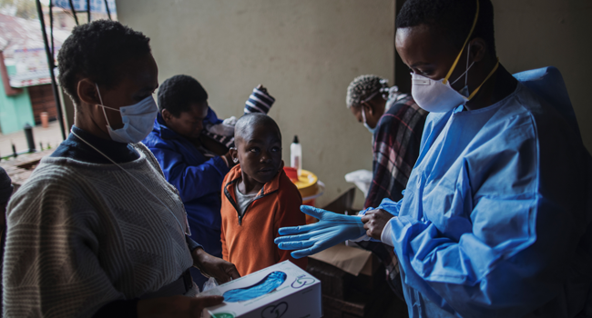 A young boy looks on as a Gauteng Health Department Official adjusts her gloves before collecting samples during a door-to-door COVID-19 coronavirus testing drive in Yeoville, Johannesburg, on April 3, 2020. MARCO LONGARI / AFP