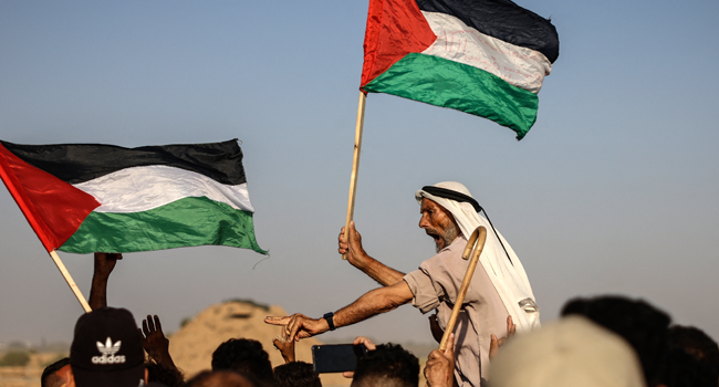 An elderly Palestinian man raises a national flag as youths shout slogans during a protest along the border fence, east of Khan Yunis in the southern Gaza Strip, on August 25, 2021. MAHMUD HAMS / AFP