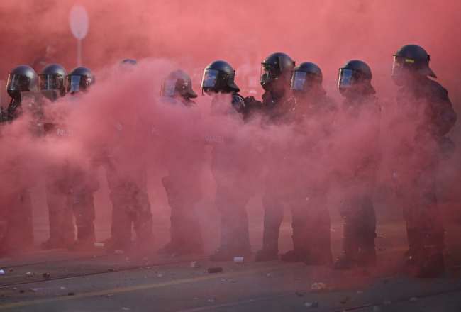 Police officers operate in riot gear with shields and rubber bullet guns amid a cloud of red smoke from a smoke bomb during a rally of a "No-G7" coalition of over 60 associations, unions and left-wing groups "against fascism and imperialism”, a day ahead of the summit's start, in Geneva on June 14, 2026. (Photo by FABRICE COFFRINI / AFP)
